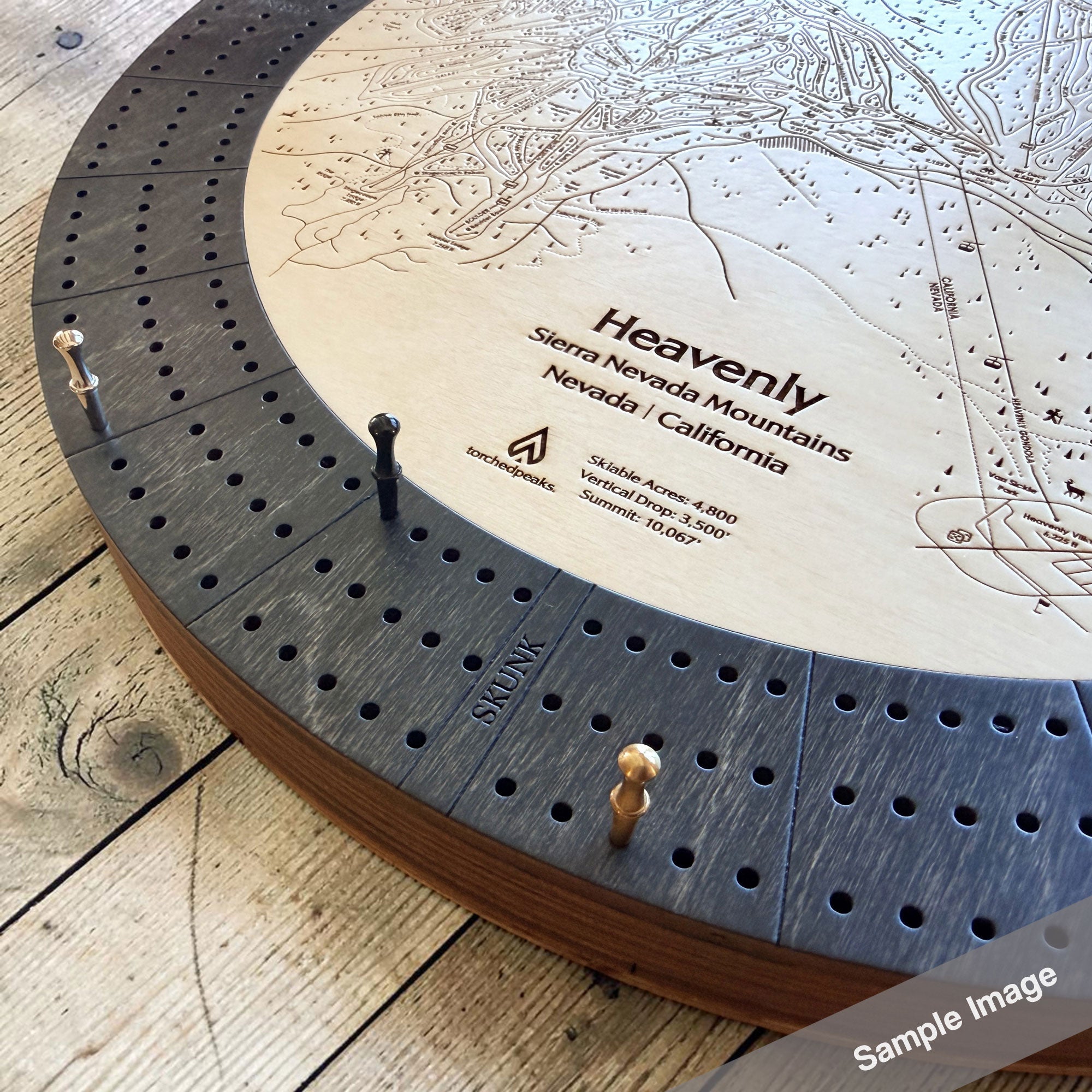 A circular wooden cribbage board game featuring an engraved ski map with dark gray cribbage peg holes around the edge and small wooden and metal pawns on a rustic wood floor.