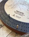A circular wooden cribbage board game featuring an engraved ski map with dark gray cribbage peg holes around the edge and small wooden and metal pawns on a rustic wood floor.