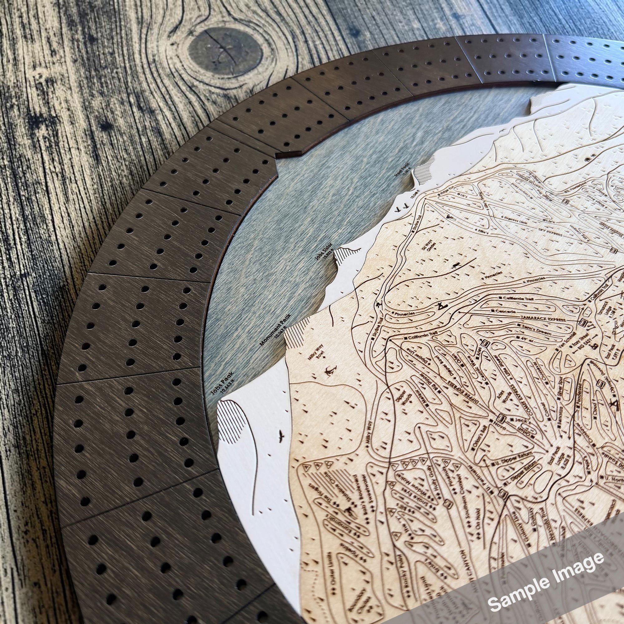 Close-up of a circular wooden cribbage board game with layered ski trail map details and a dark wood segmented playing track, photographed on a rustic wood surface. 