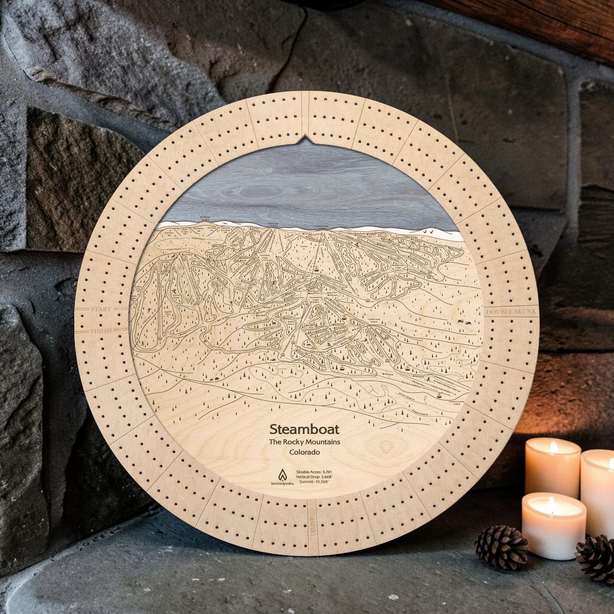 Birch Steamboat ski trail map wooden cribbage board displayed by a stone fireplace with candles and pinecones.