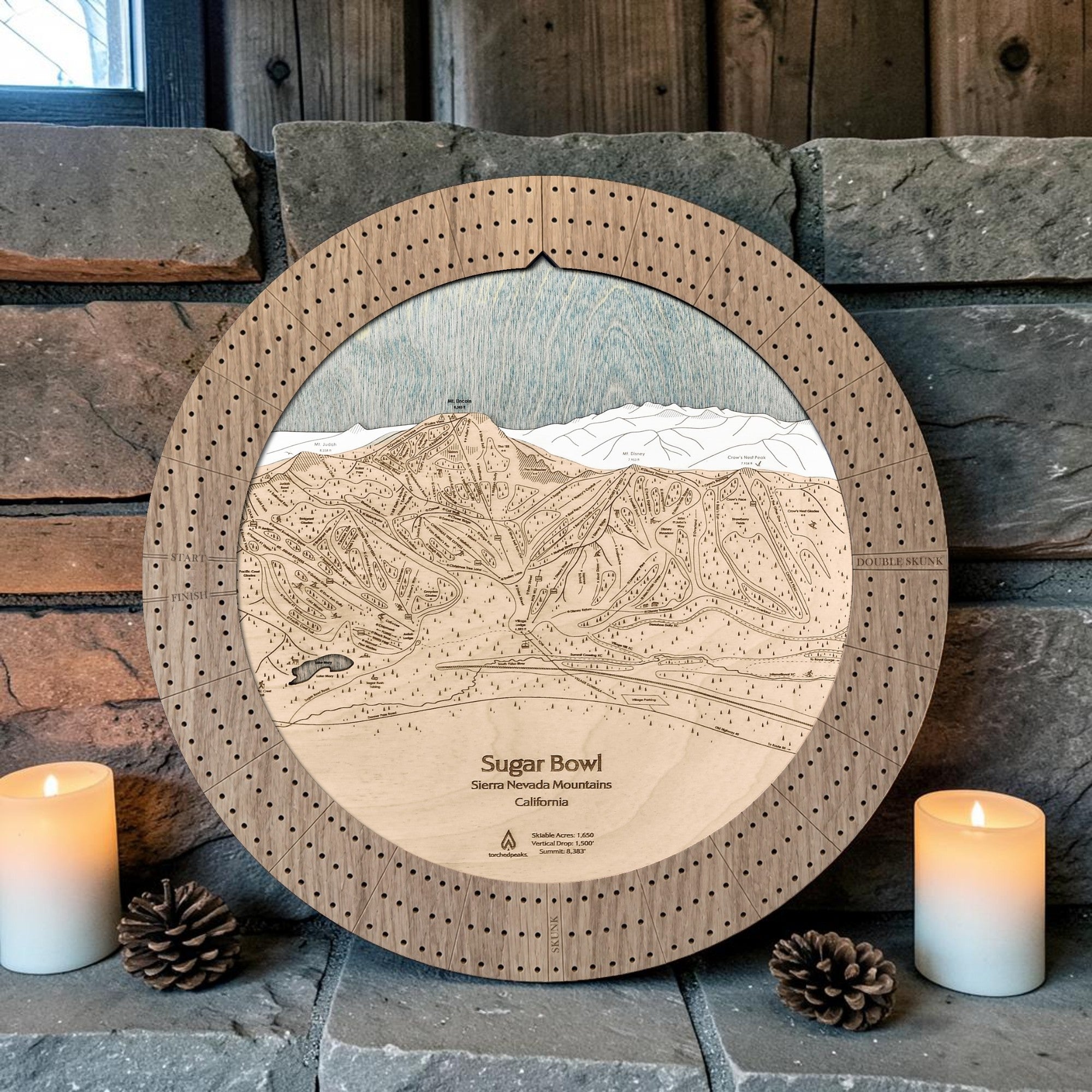 Sugar Bowl cribbage board in Weathered Timber beside a stone fireplace with candles and pinecones.