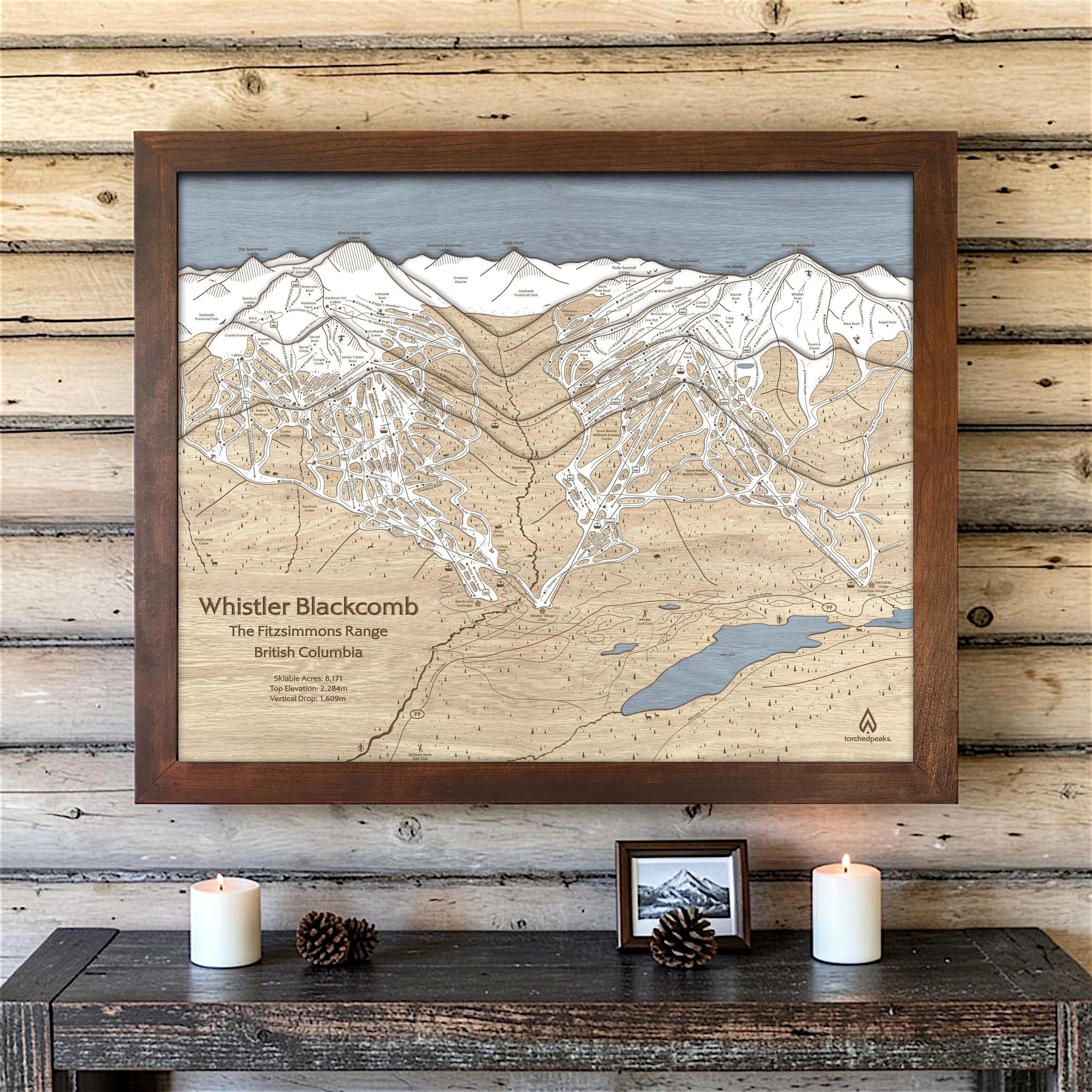 Rustic Mocha-framed Whistler Blackcomb wood ski map on a light cabin wall above candles, pinecones and a small photograph.