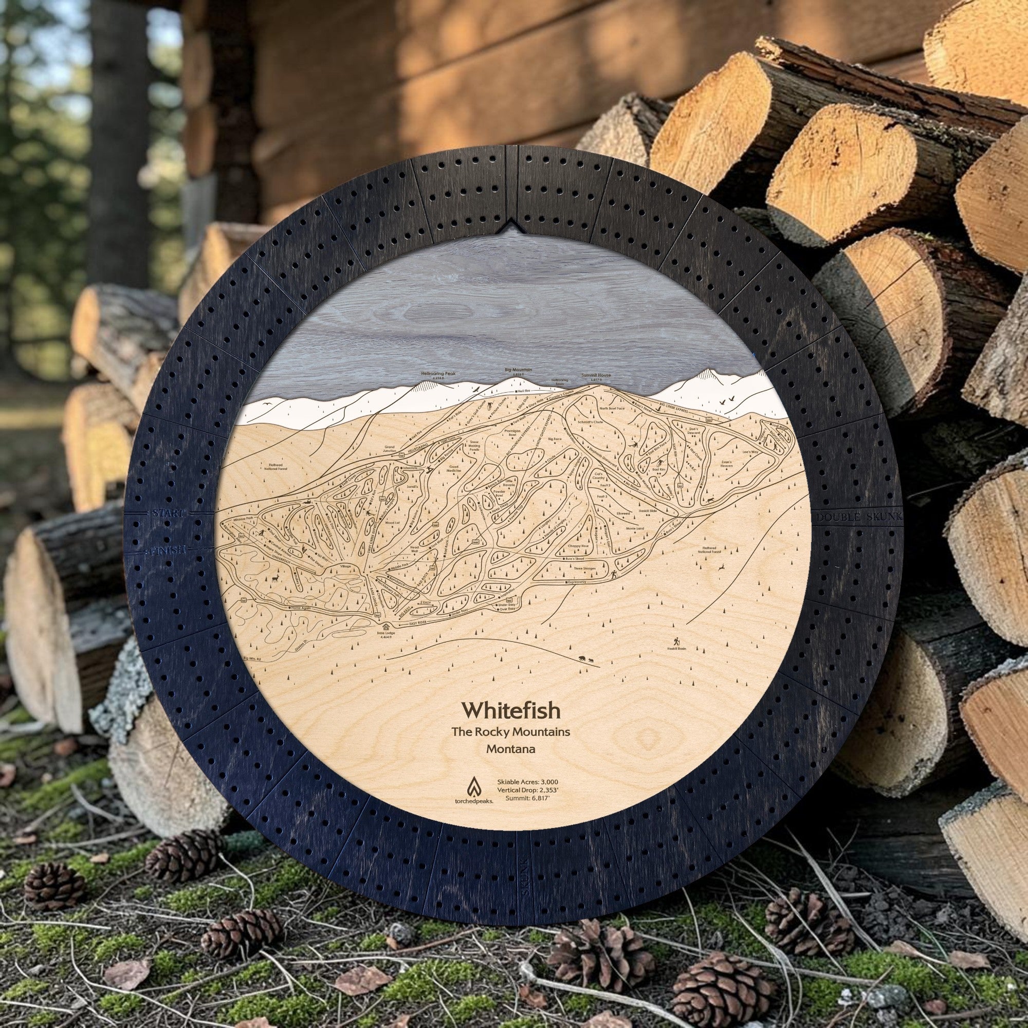 Matte Onyx Whitefish ski map cribbage board staged beside stacked firewood in a mountain cabin setting.