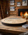 A rustic wooden ski map board game set displayed on a wooden table in a cozy cabin living room with a burning fireplace, skis leaning against the wall, and framed ski photos on the wood-paneled wall.